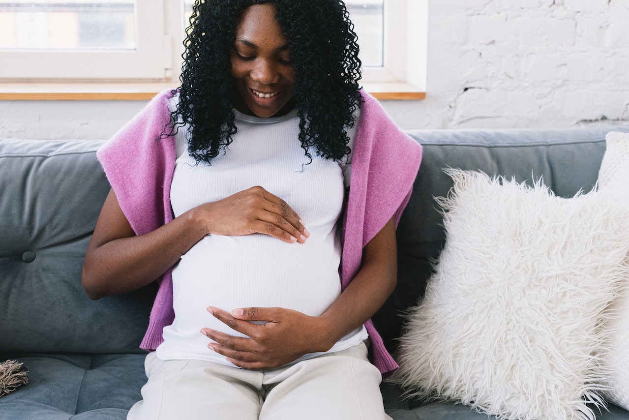 A Black pregnant woman sitting on a couch looks down, smiling and cradling her stomach.