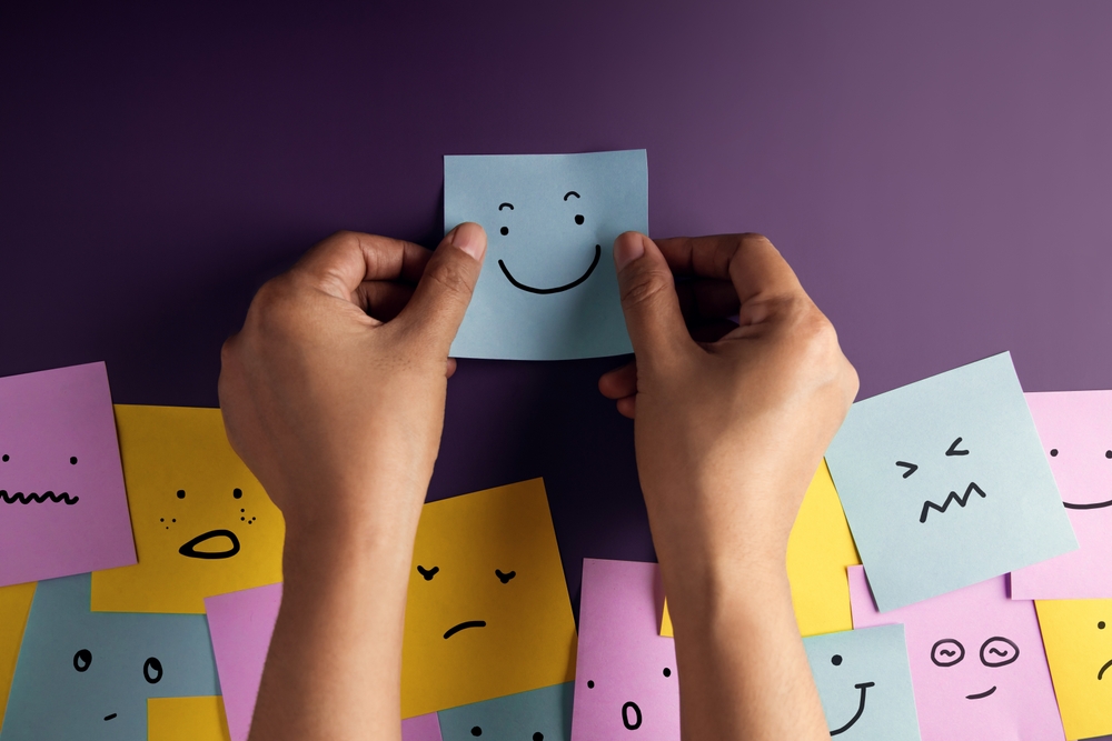 Pair of hands placing a blue sticky note with a smiley face drawing on a purple background. There are sticky notes with different colors and drawings of facial expressions in the background.