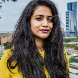 Sadiya Muqueeth headshot. Sadiya has long brown hair and is wearing a yellow sweater. A city skyline is behind her.