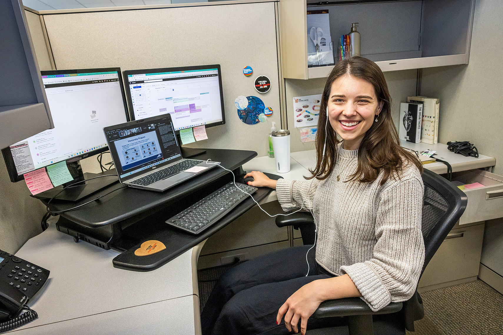 A smiling woman sits at a desk. There are two desktop computer screens and one laptop at her workstation.