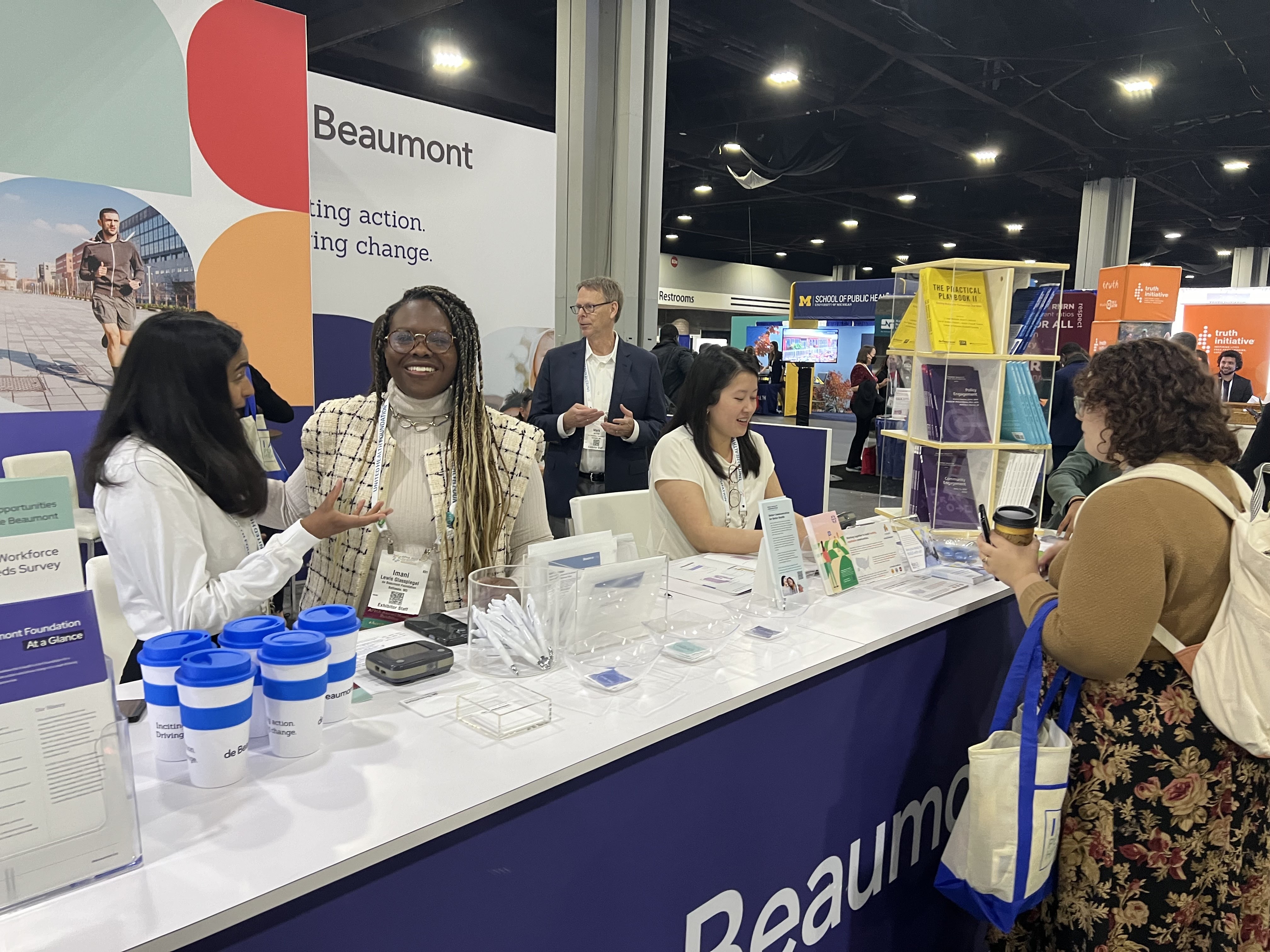 de Beaumont staff at their Annual Meeting booth. Several staff stand behind a table, smiling and talking to each other and an APHA attendee.