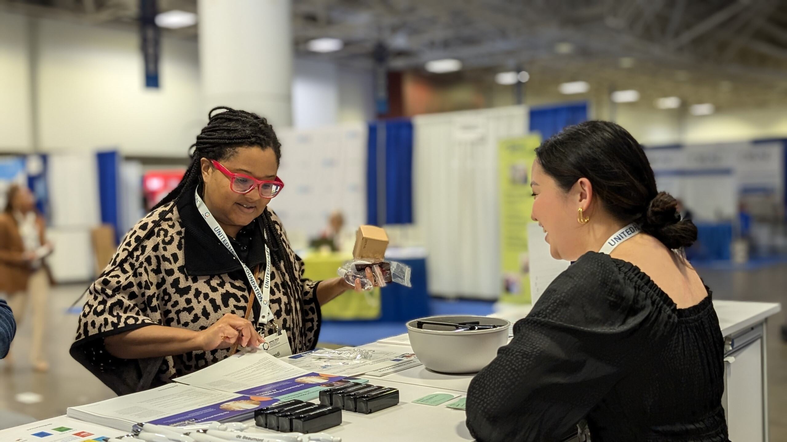 Two people at the de Beaumont booth in the APHA 2024 expo hall. One woman stands in front of the booth table, which is covered in handouts and giveaway items. A de Beaumont staff member stands behind the table.