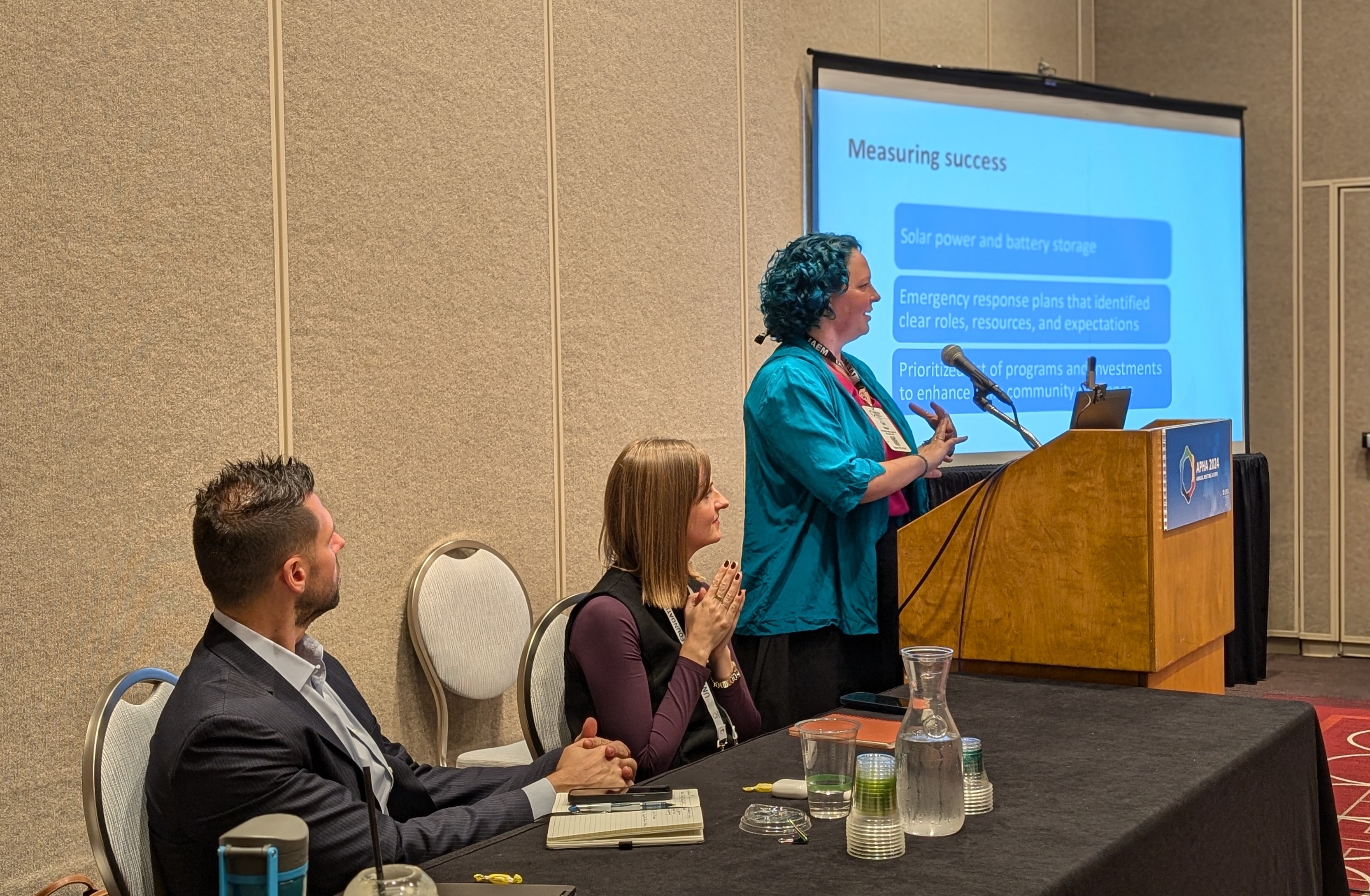 Two people sit at a table facing a person standing to present at a lectern.