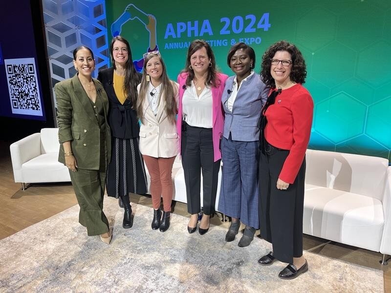 Six smiling women stand in a row on a stage in front of a screen with the APHA 2024 logo.