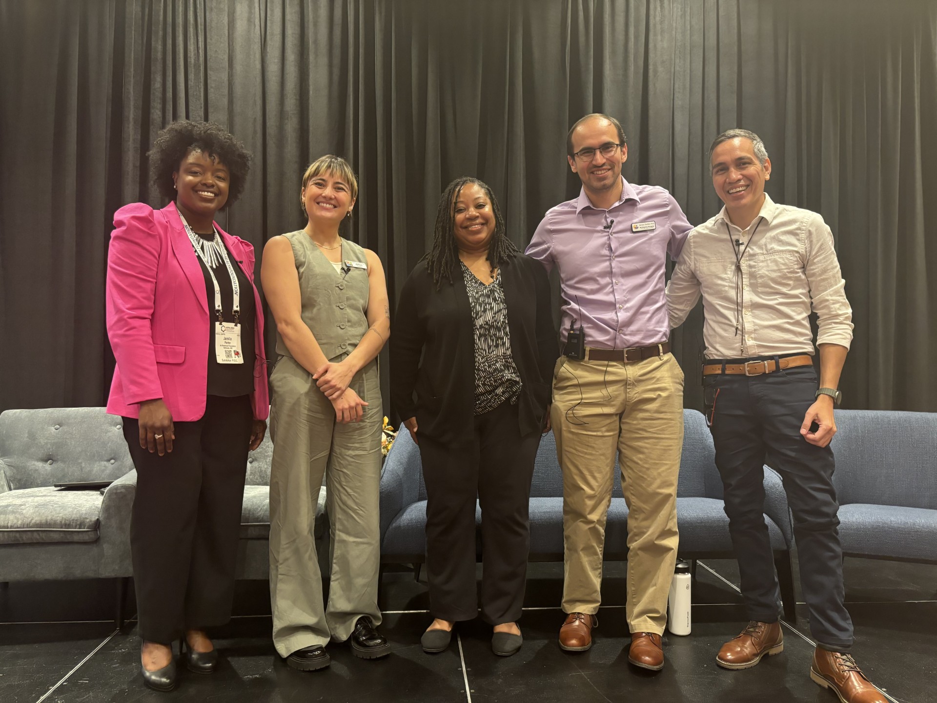 Five smiling people stand in a row. They are standing in front of a black curtain on a conference stage.