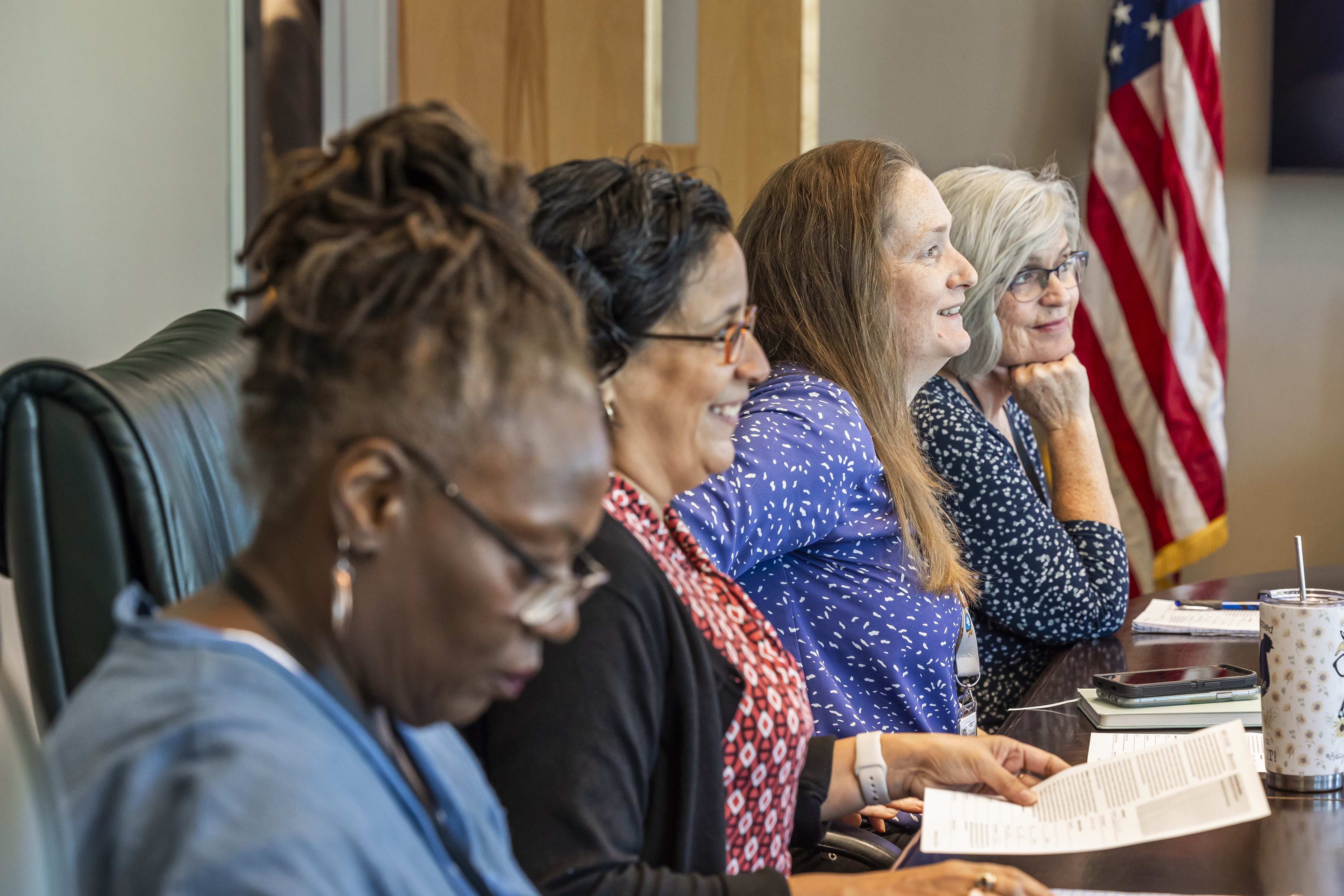 Four women in a row sit at a table. Three women are looking up and one is looking down.