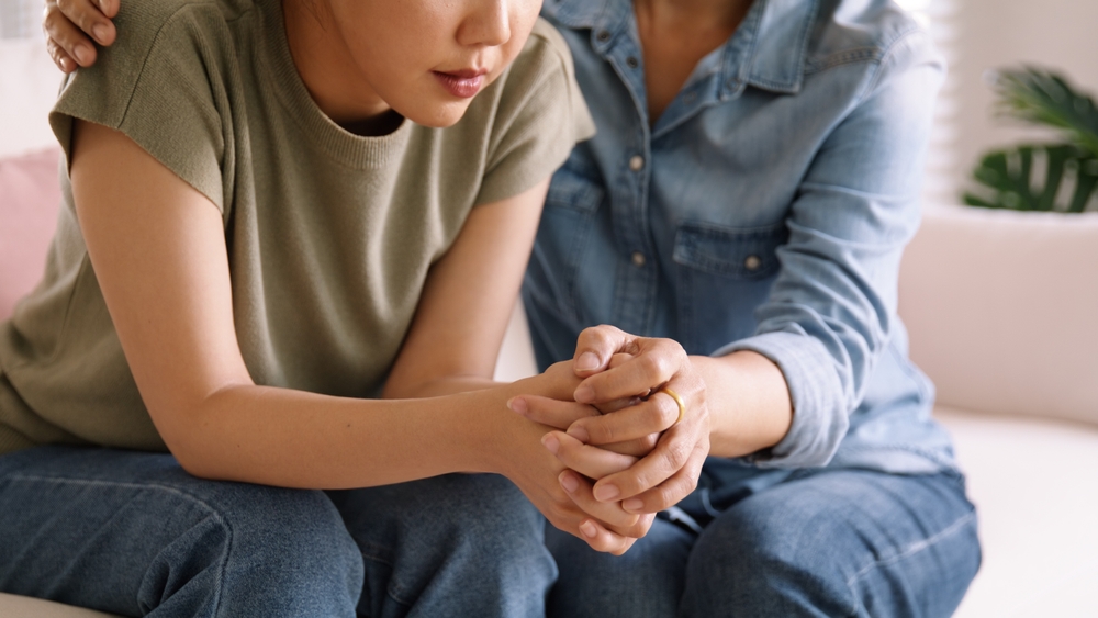 Two people visible from the neck down sit on a couch. The person on the right puts her left hand on the other woman's interlaced fingers and her right hand on the woman's shoulder.