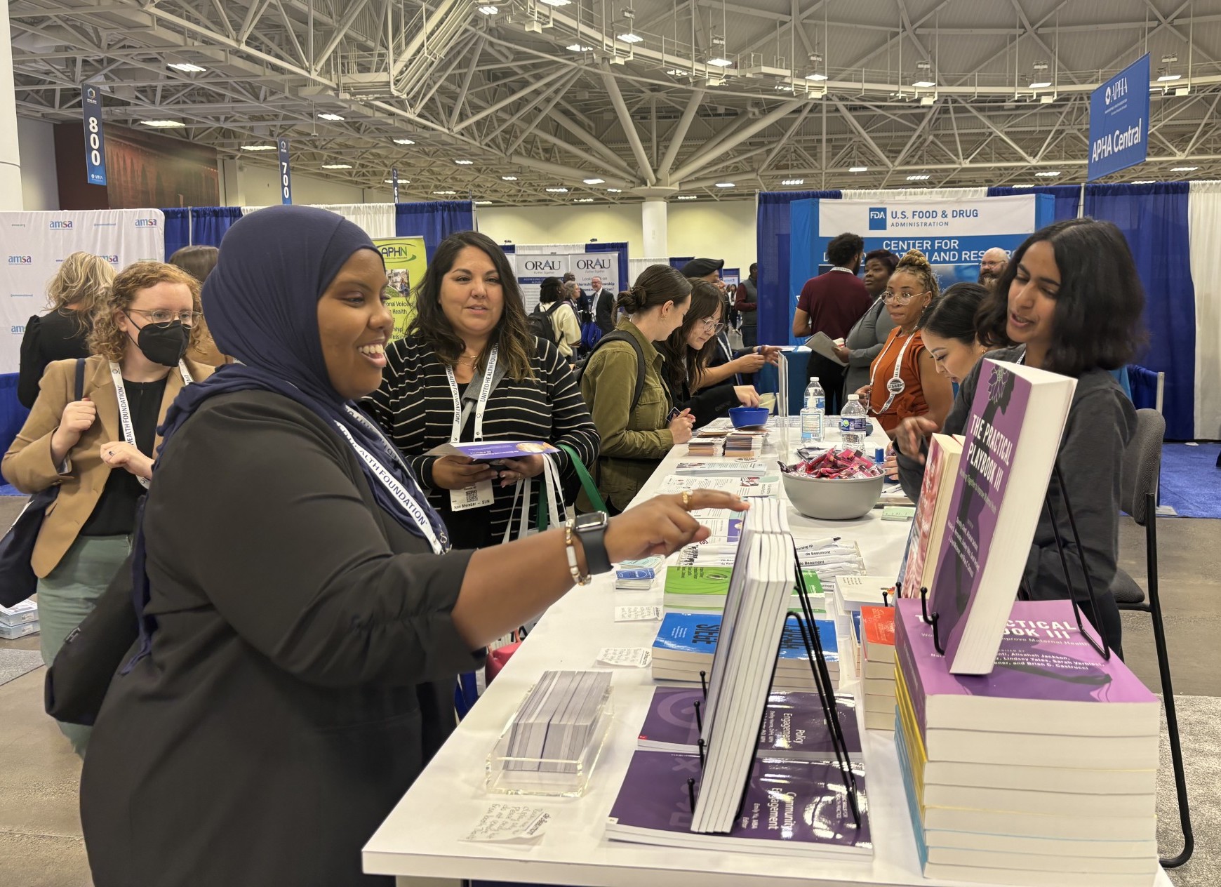 APHA 2024 attendees browse the offerings on the de Beaumont booth table. de Beaumont staff stand behind the table.