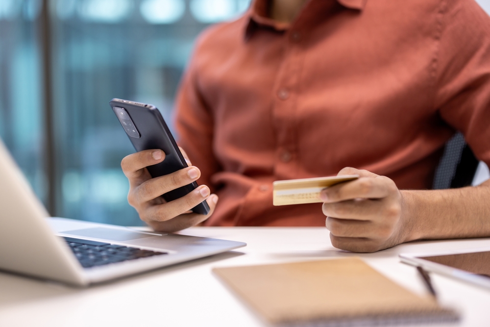 Image of a person from the neck down sitting in front of an open laptop. He is holding a credit card in one hand and a cell phone in the other.