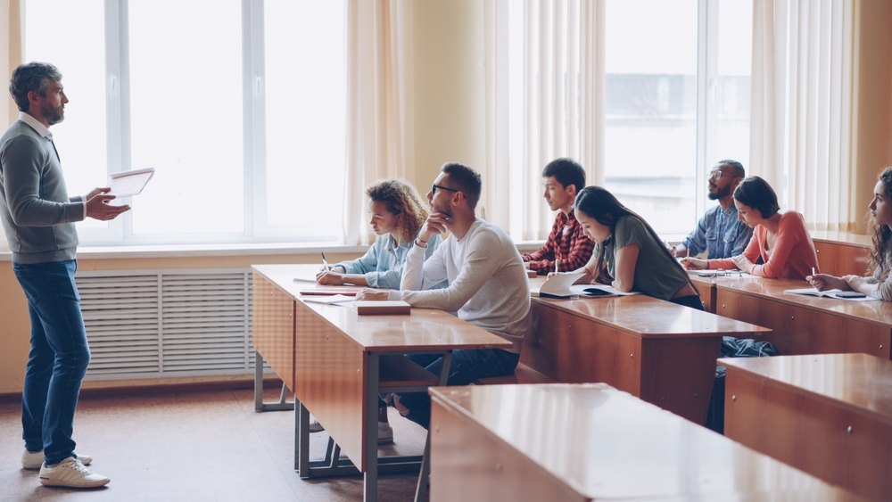 A teacher speaks to a class of students sitting in rows of desks.