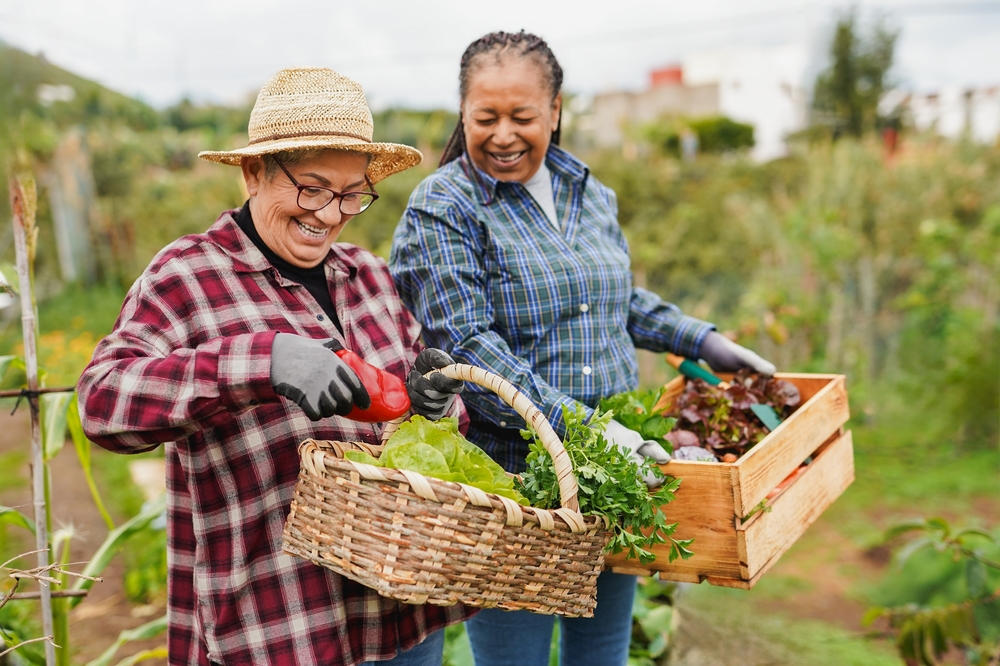Two smiling people in a field. One person carries a basket of produce and the other carries a box of produce.